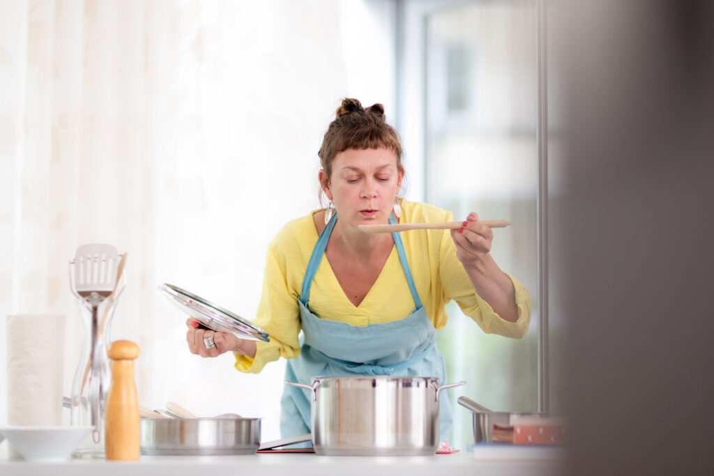 Inga beugt sich über einen Topf und pustet auf einen großen Holzlöffel, den sie in der Hand hält.